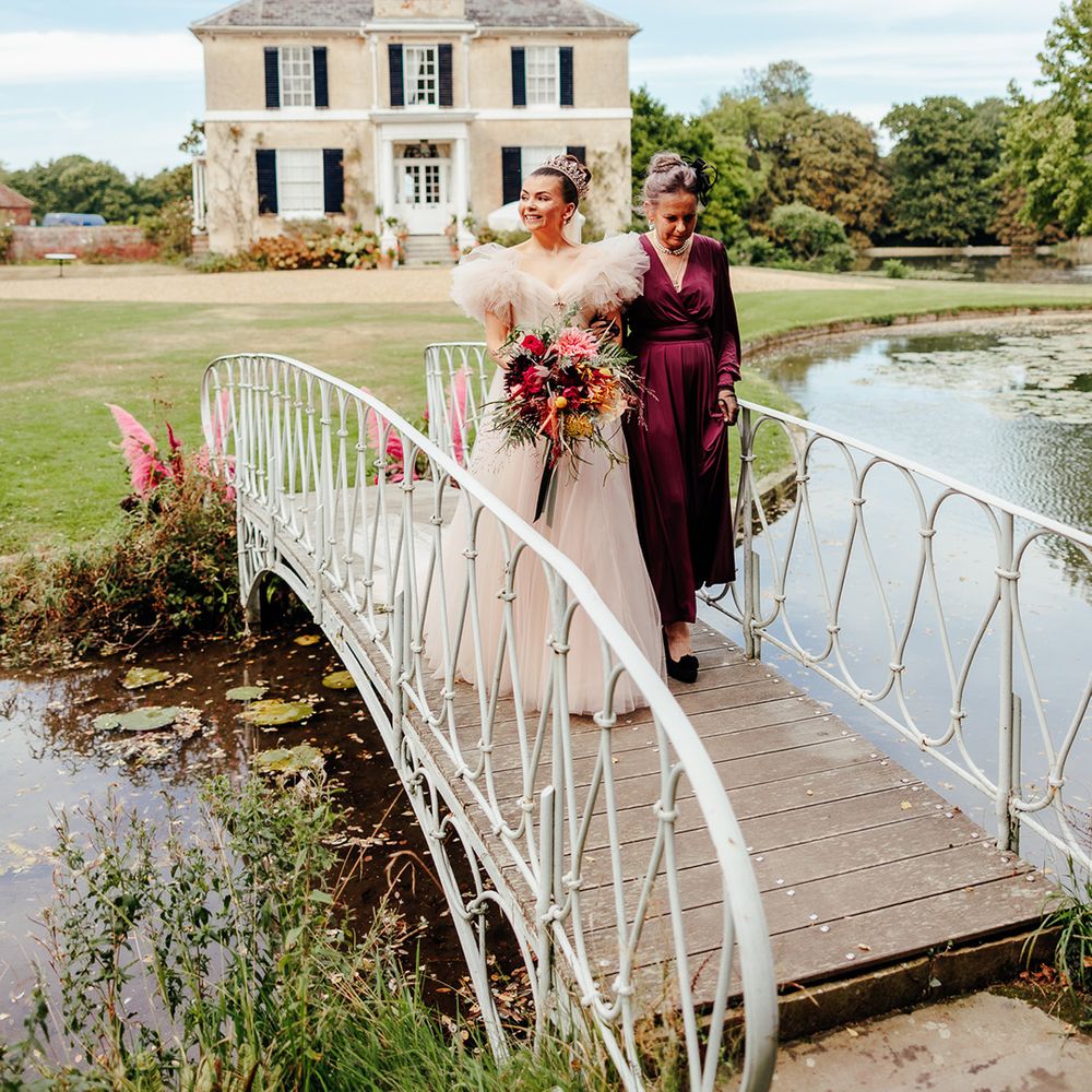 Mother of the bride and bride walk across bridge to outdoor wedding ceremony space