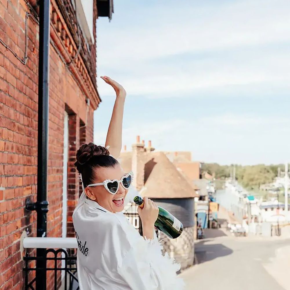 Bride drinking champagne, wearing white robe and heart glasses for hen do