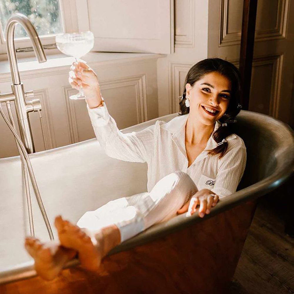 Bride in white satin bridal pyjamas having a glass of wine in the bath