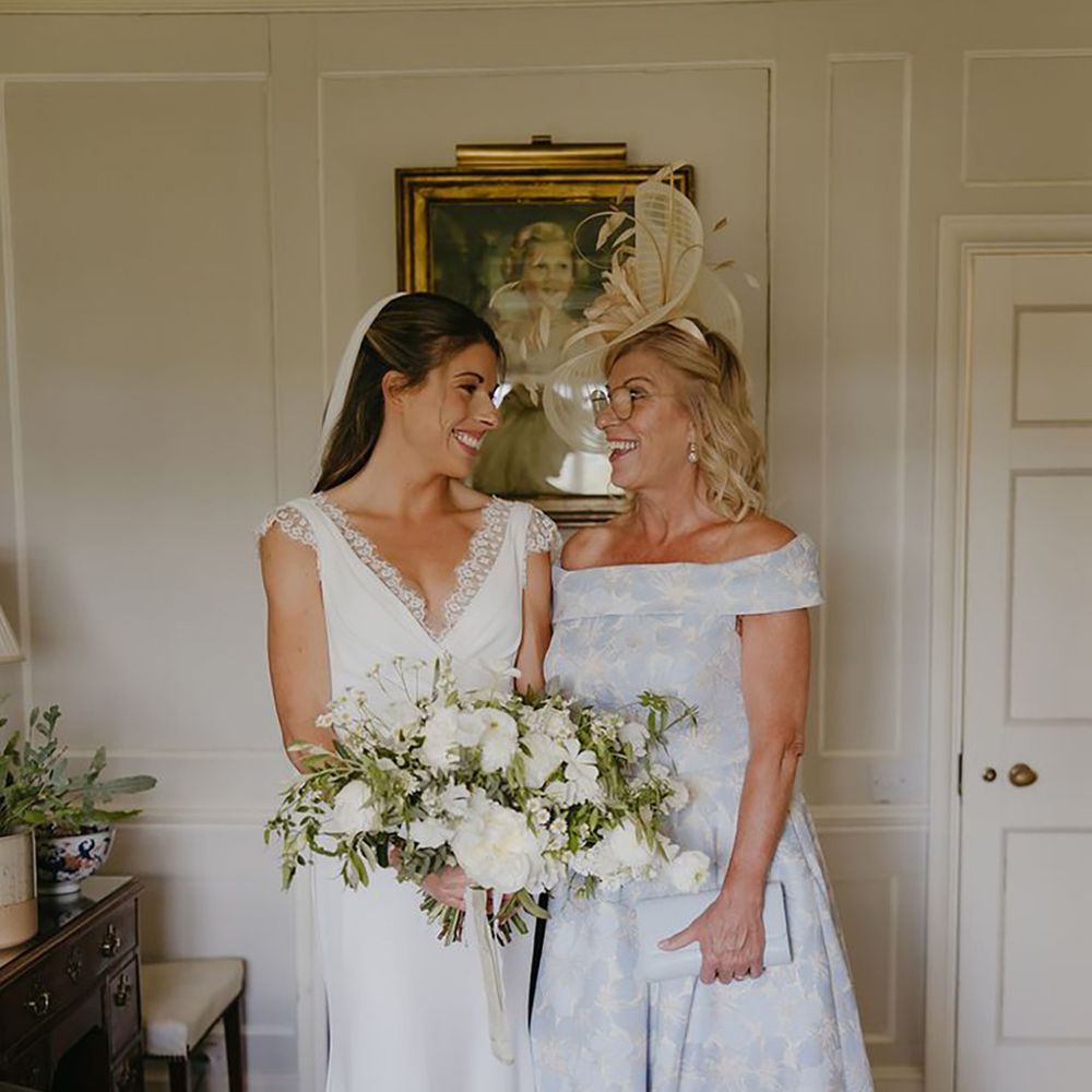 Bride in lace wedding dress with white bouquet smiling at mother of the bride in floral dress