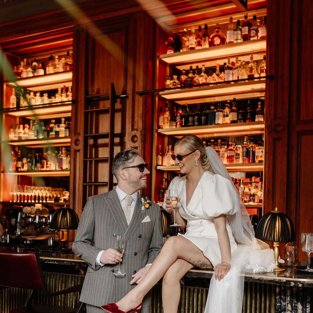bride-wearing-red-wedding-shoes-sits-on-wedding-bar-next-to-groom-in-grey-suit.