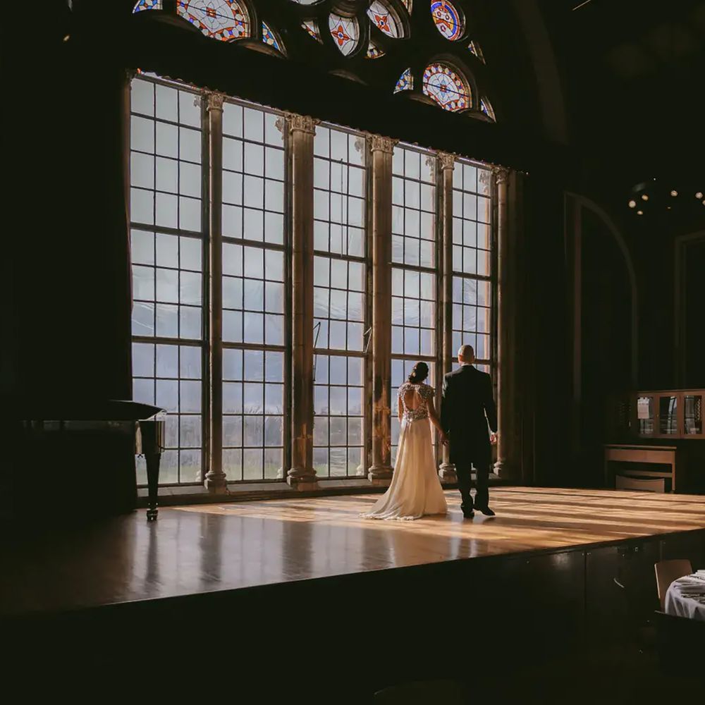 Couple standing by large windows of the Barry Buildings Great Hall at sustainable wedding venue, Dulwich College