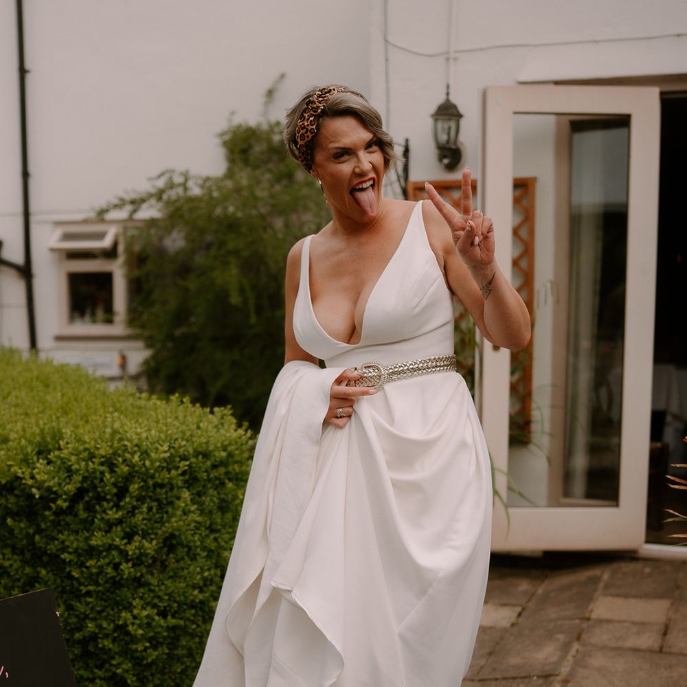 bride-in-wedding-dress-with-gold-belt-makes-peace-sign-and-sticks-out-tongue