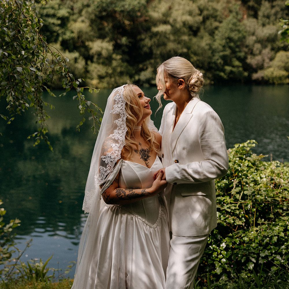 bride-in-embroidered-edge-wedding-veil-smiles-up-at-bride-in-suit-in-front-of-lake