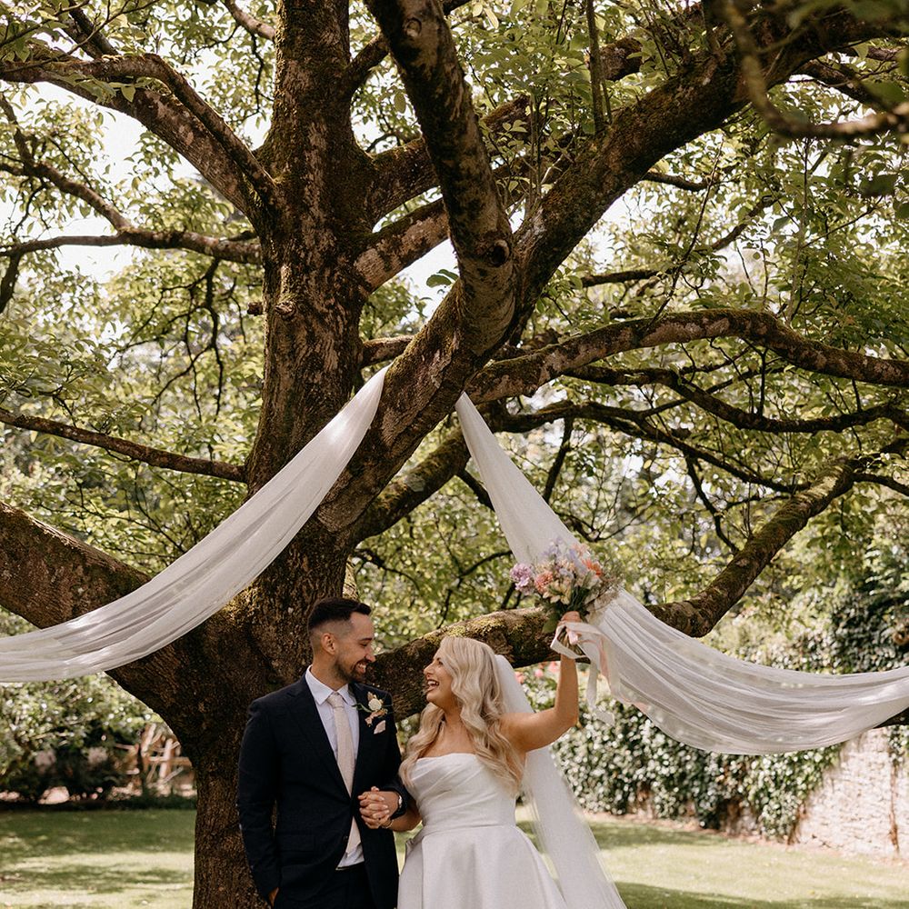 couple-celebrating-under-tree-at-outdoor-wedding-with-fabric-drapes-pastel-wildflower-bouquet