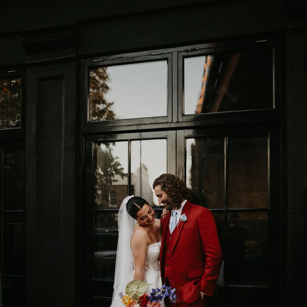 groom-in-red-suit-looking-at-bride-in-strapless-wedding-dress-carrying-colourful-bouquet