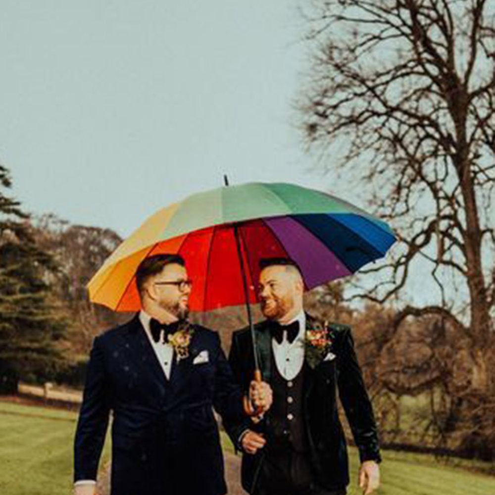 Two grooms walking under large rainbow golf umbrella
