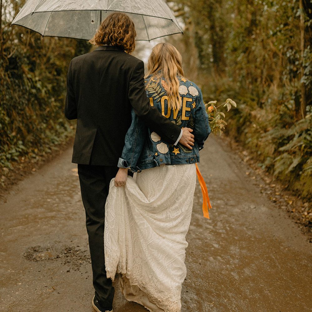 Bride in embroidered motif denim jacket walking under clear umbrella with groom 