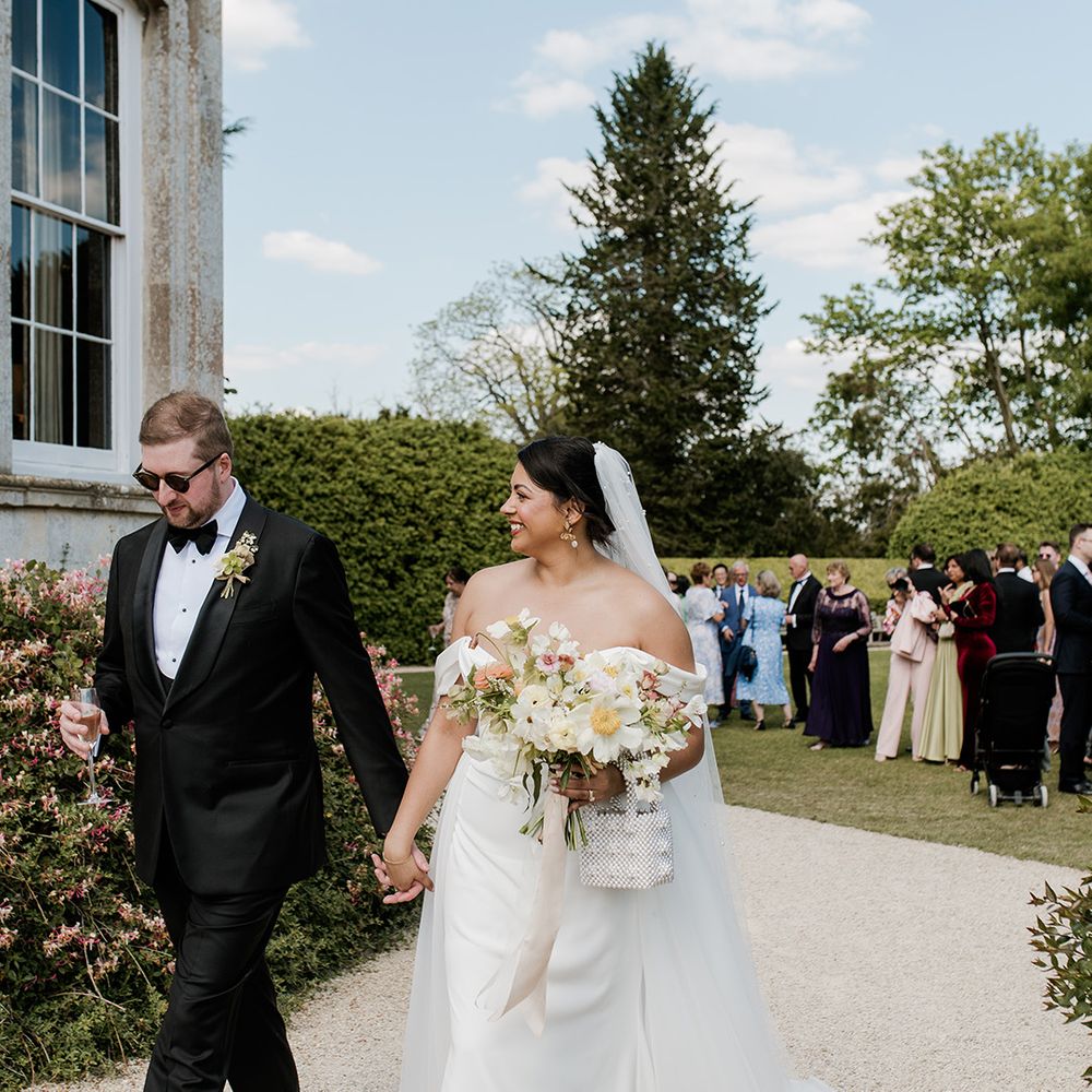 groom-in-black-tuxedo-with-sunglasses-walks-with-bride-in-off-the-shoulder-wedding-dress