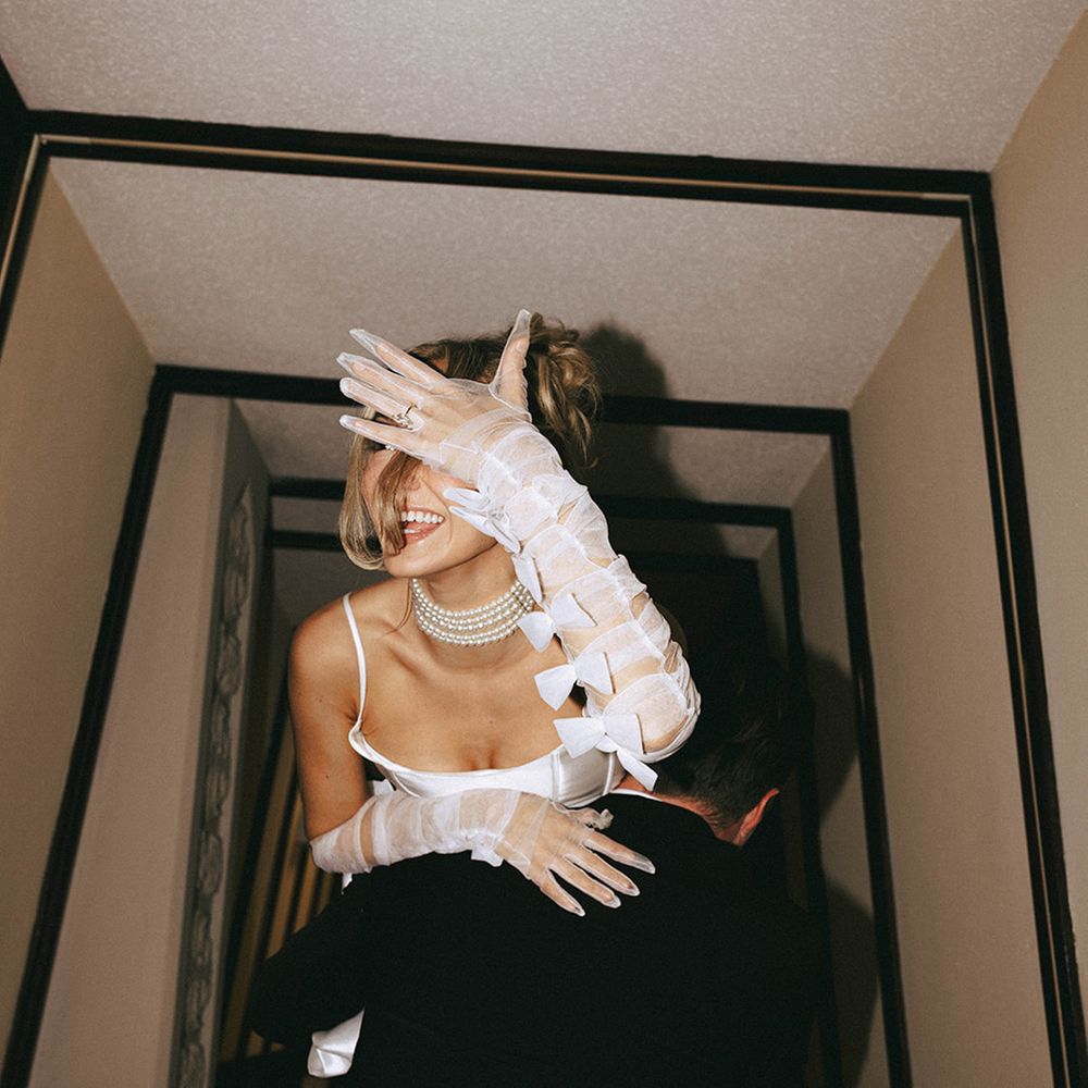 Man carries fiancee with bow bridal gloves and pearl choker showing off ring