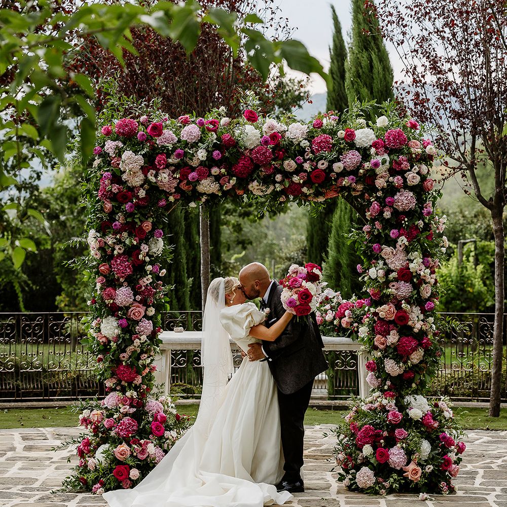 couple-kisses-under-pink-flower-arch-at-tuscany-wedding