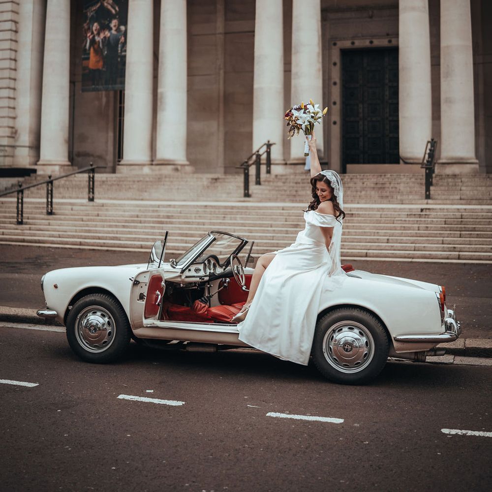 Bride sitting on the back of convertible wedding car with bouquet in the air