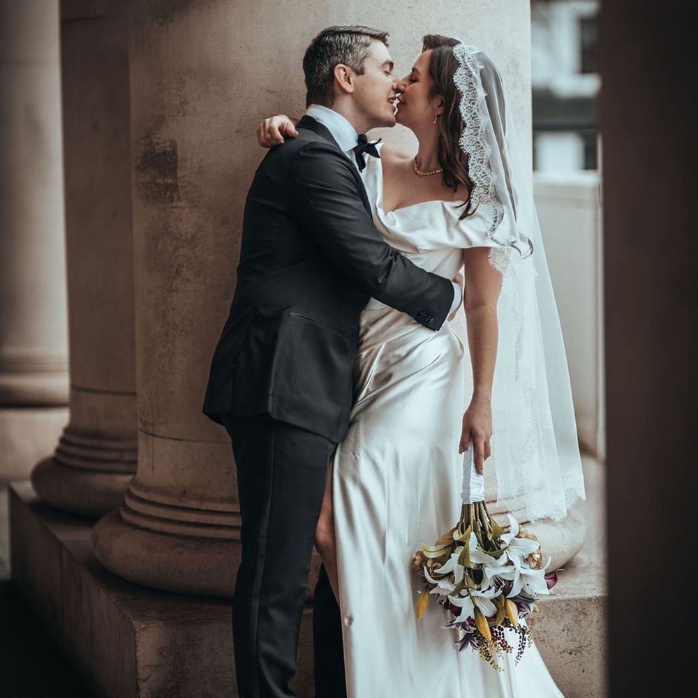 Groom kisses bride in off the shoulder wedding dress with embroidered lace edge veil