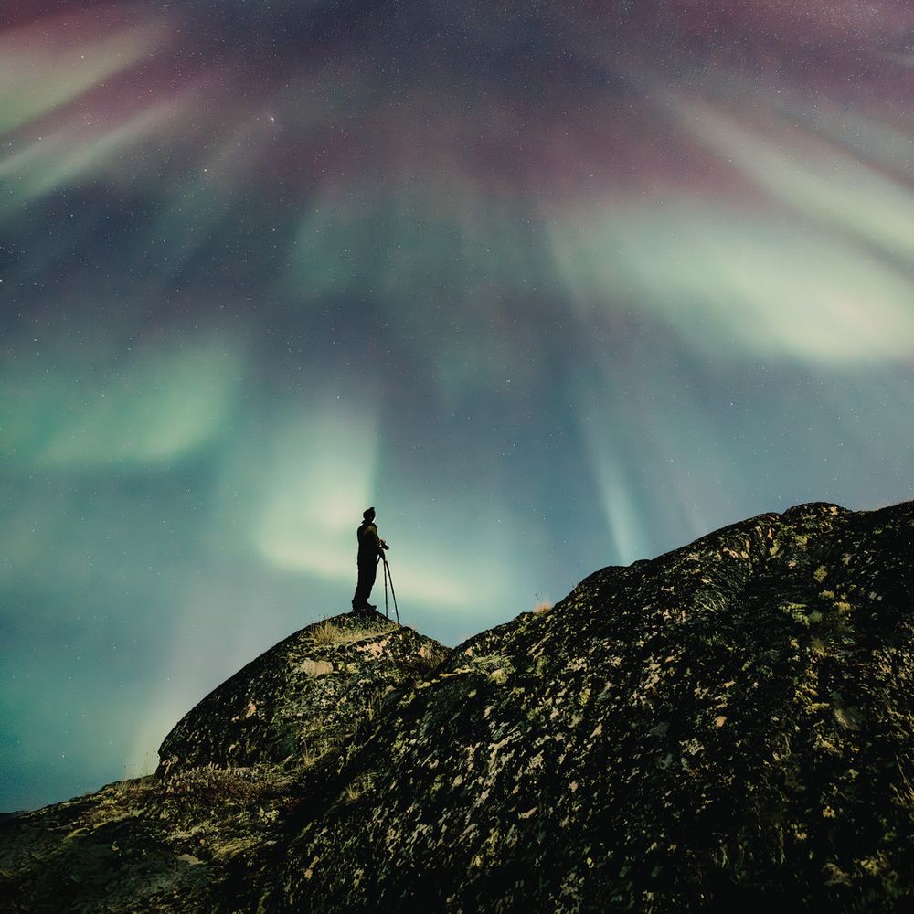 Man stands on cliff under Aurora Borealis