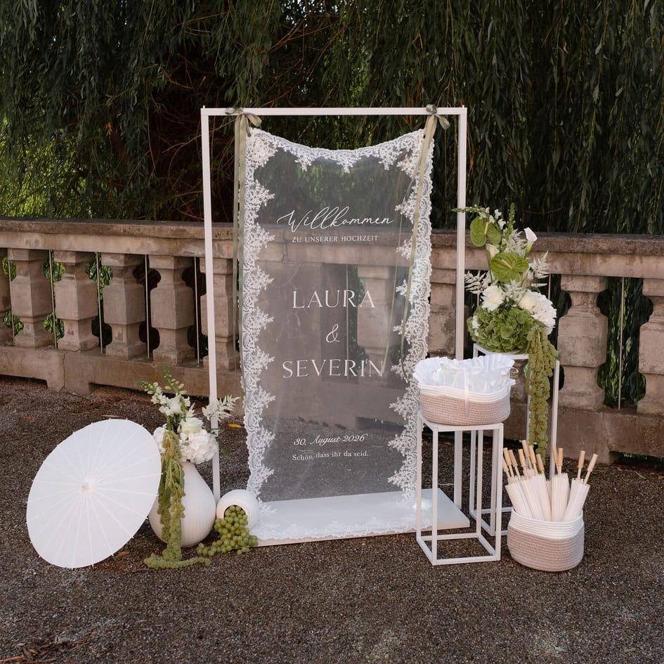 Acrylic wedding welcome sign draped in lace beside white parasols and green floral displays