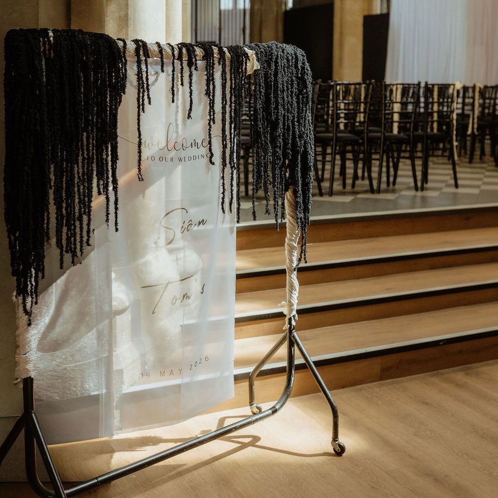 Frosted acrylic welcome sign draped with black macramé fringing in a stone church ceremony space