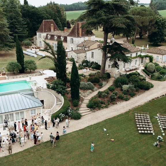 Aerial view of guests gathered by a poolside château with an outdoor ceremony set-up on the lawn