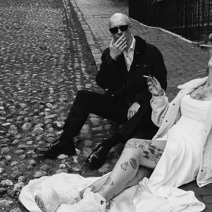 Newlyweds in sunglasses sit on cobbled ground, bride in white dress and trainers with tattoos