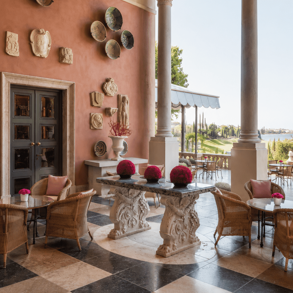 Elegant Mediterranean-style hotel terrace with vaulted arches, marble columns, wicker lounge seating, decorative ceramic wall displays, and panoramic views of landscaped gardens, a golf course, and a lake