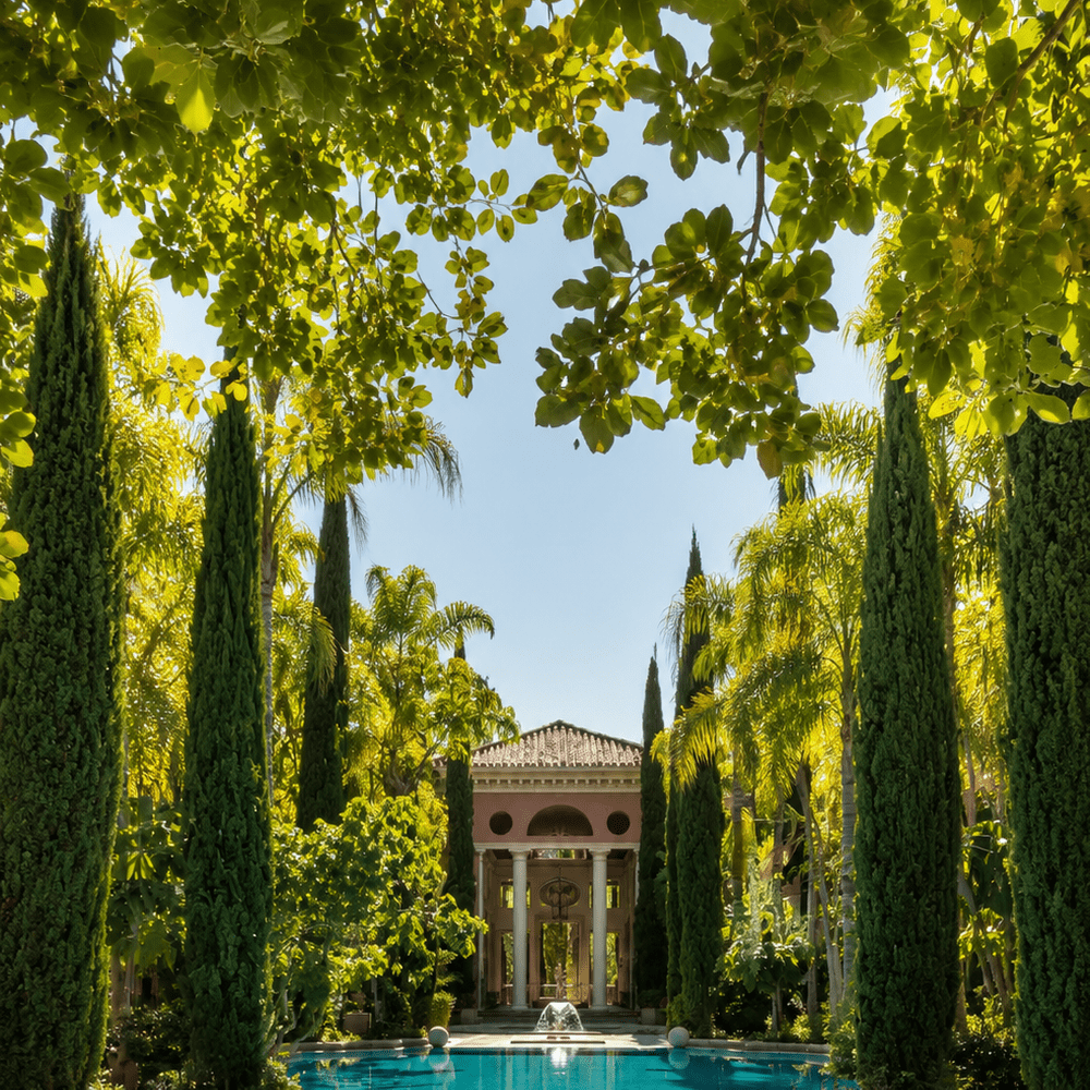 luxurious outdoor swimming pool surrounded by tall cypress and palm trees, leading to an elegant Mediterranean-style villa with classical columns and lush landscaped gardens under a bright blue sky