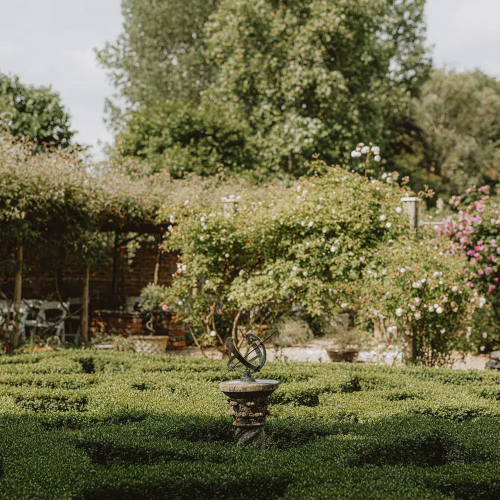 Armillary sundial at the centre of a clipped box hedge parterre with climbing roses behind