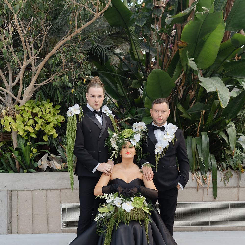 Bride in black off-shoulder gown with orchid crown seated between two men in black tuxedos amid tropical foliage