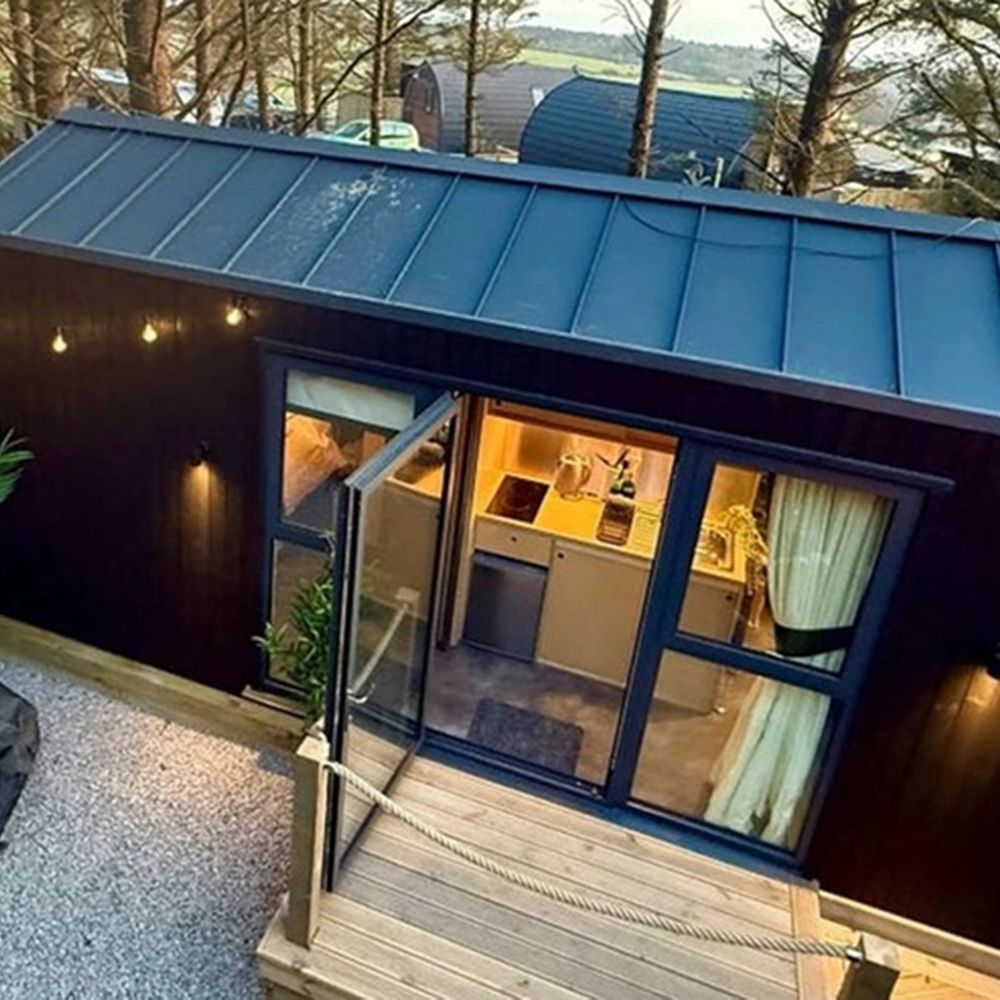 Black-clad woodland cabin with open glass doors, festoon lights and grey beanbags on a gravel terrace