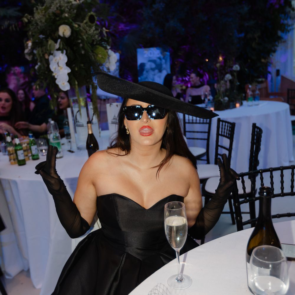 Guest in dramatic black gown, wide-brim hat and gloves poses at an evening reception table
