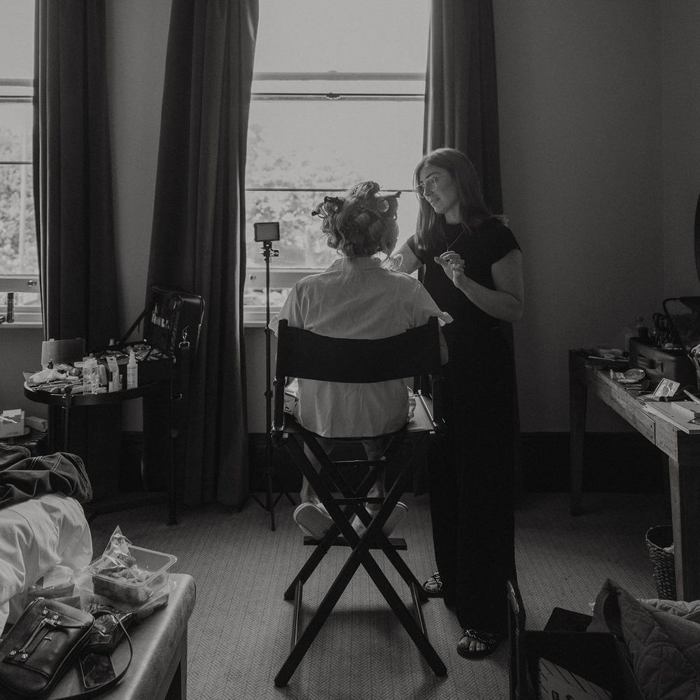 Bride in curlers has her make-up applied by an artist in a bright window-lit room