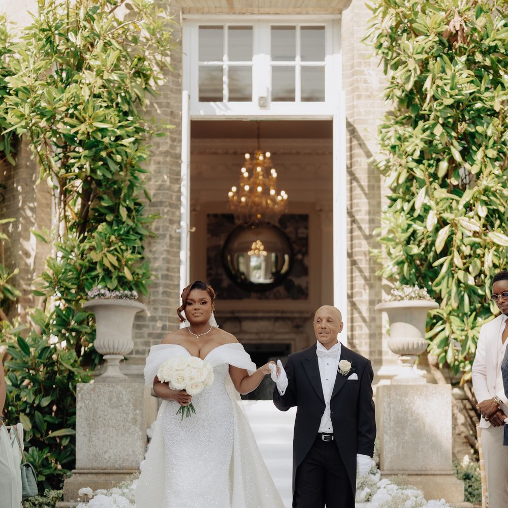 Bride in off-shoulder gown walks down white aisle lined with blooms beside a smartly dressed man