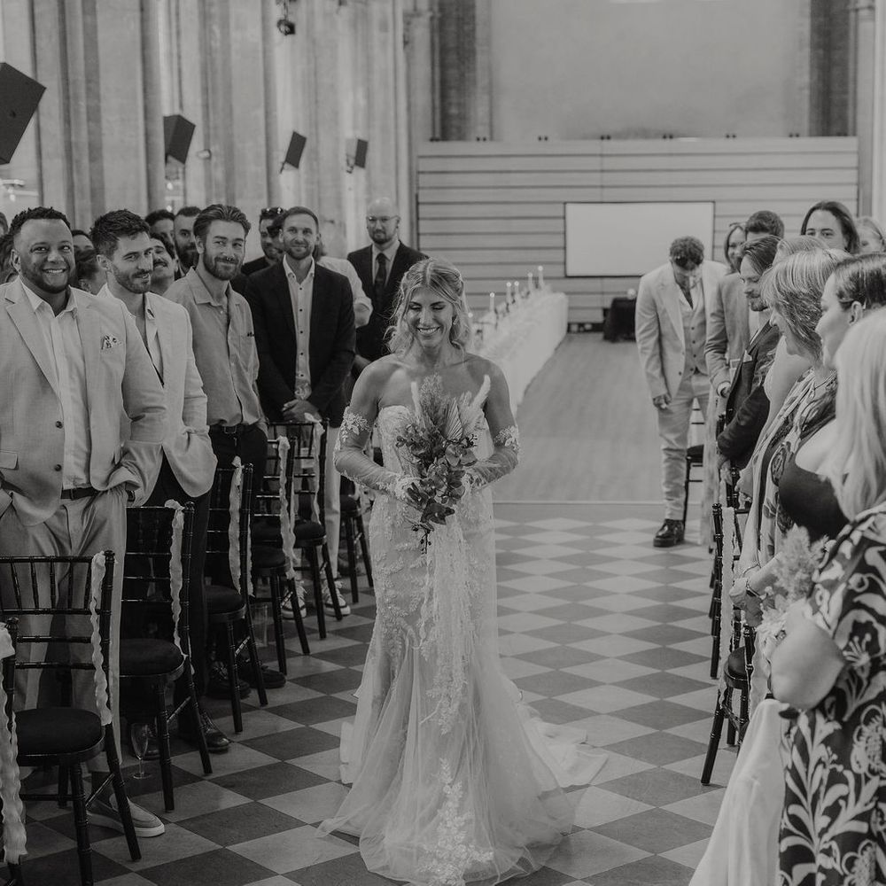 Smiling bride walks down a checkerboard aisle holding a bouquet as guests look on