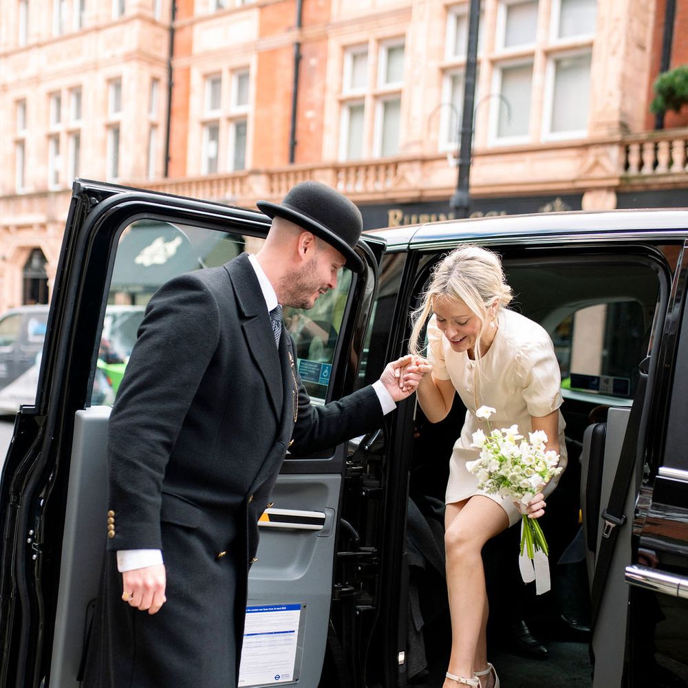 Bride in a short dress steps from a black cab as a doorman offers his hand