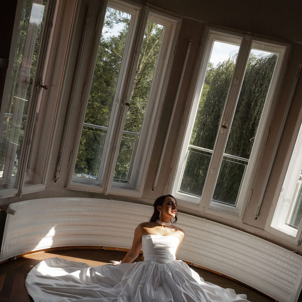 Bride in strapless ball gown seated on parquet floor by tall bay windows in soft light