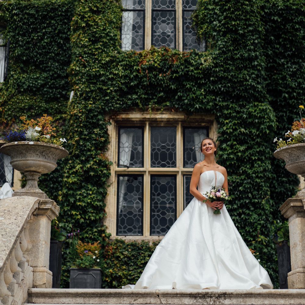 Bride in a strapless ballgown holding a bouquet on stone steps before an ivy-clad manor