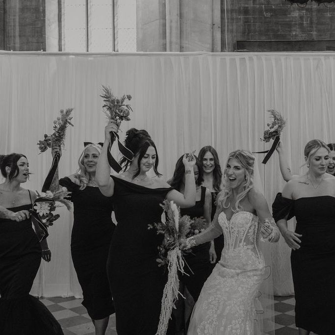 Bride and bridesmaids in black dresses laughing and holding bouquets aloft