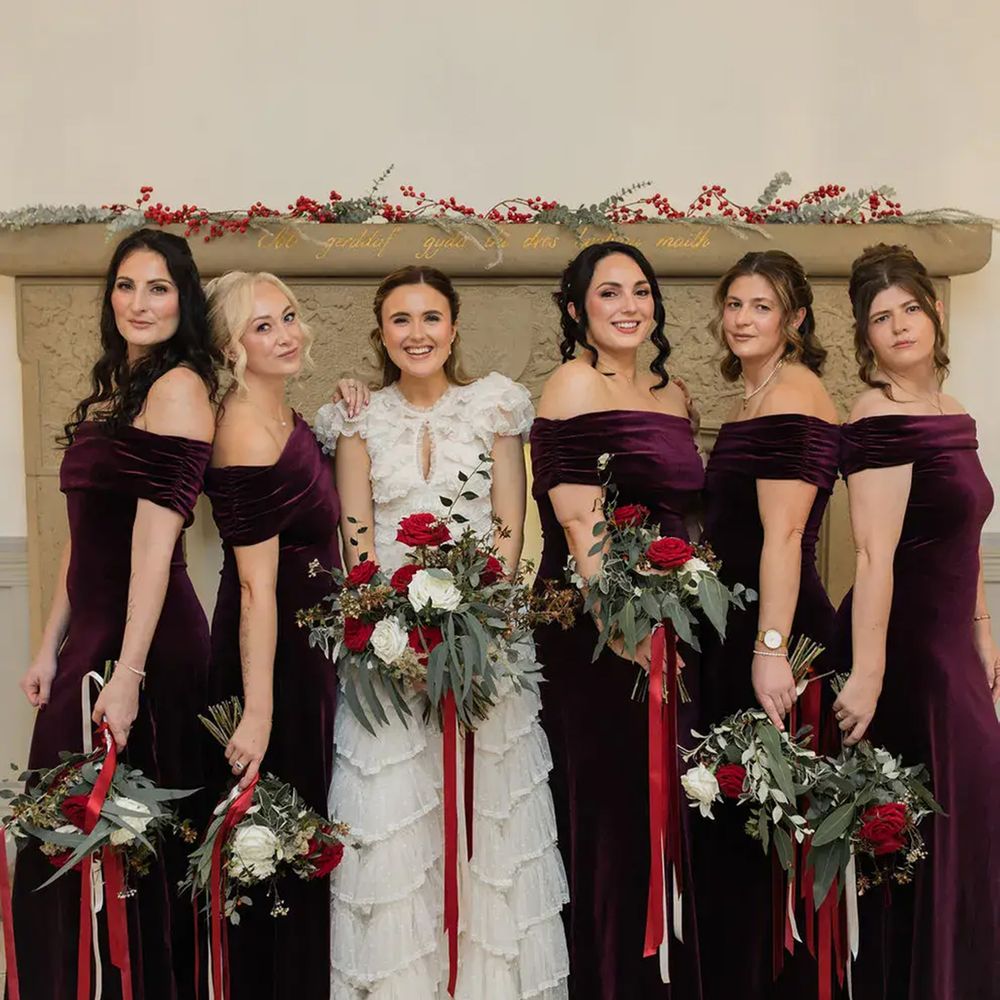 Bride in tiered lace gown with five bridesmaids in burgundy velvet dresses holding red and white bouquets