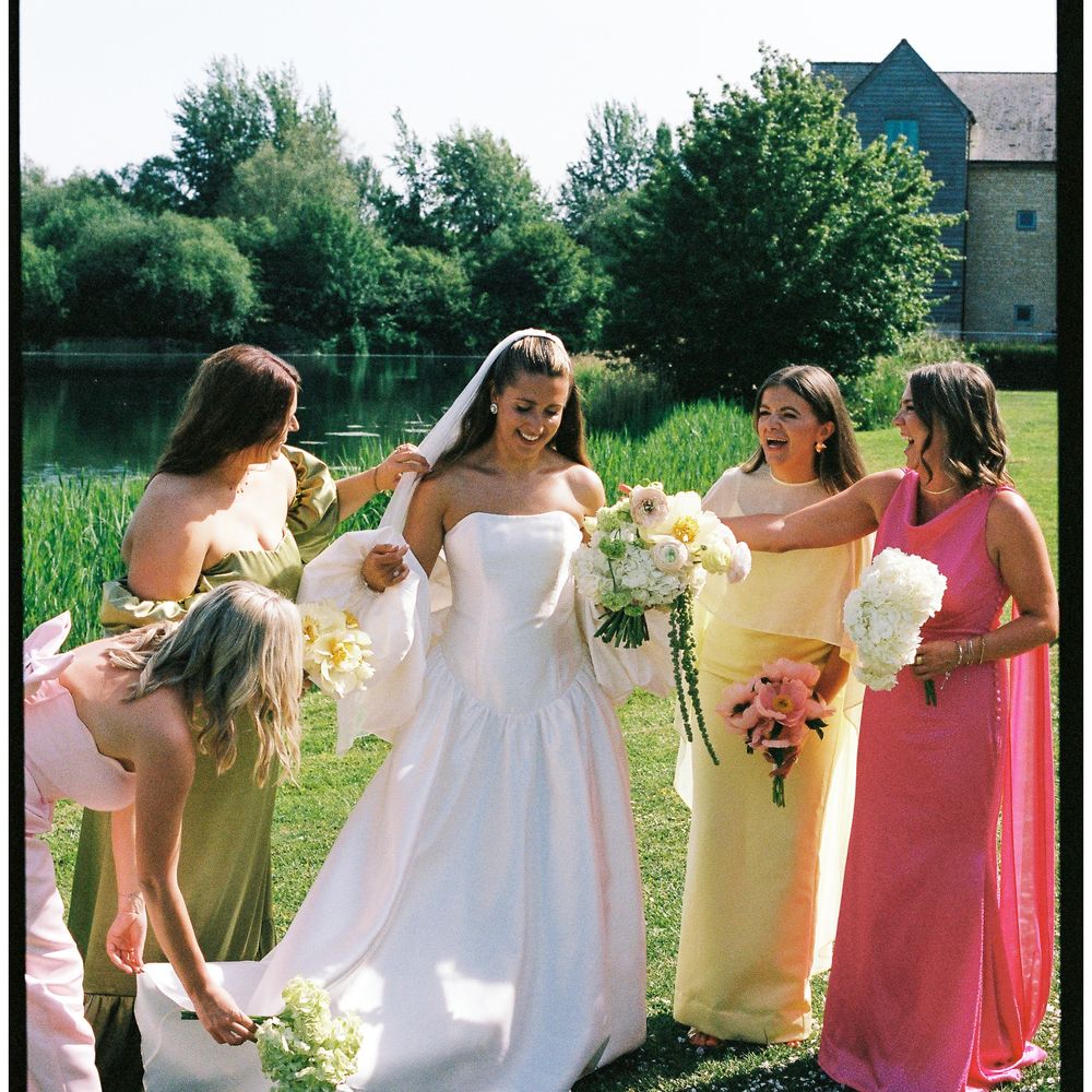 Bride laughing with bridesmaids in green, yellow and pink dresses by a lakeside lawn