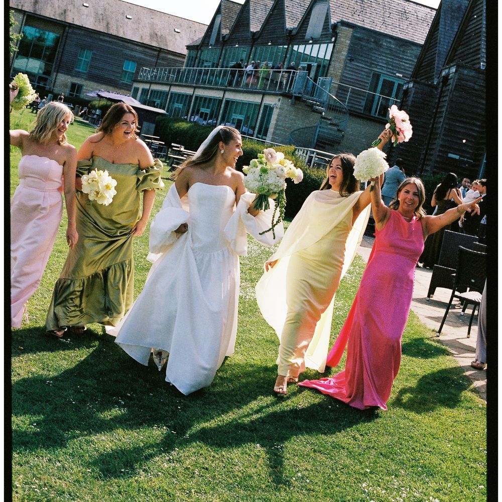 Bride and bridesmaids in colourful gowns walking across a sunlit lawn holding bouquets