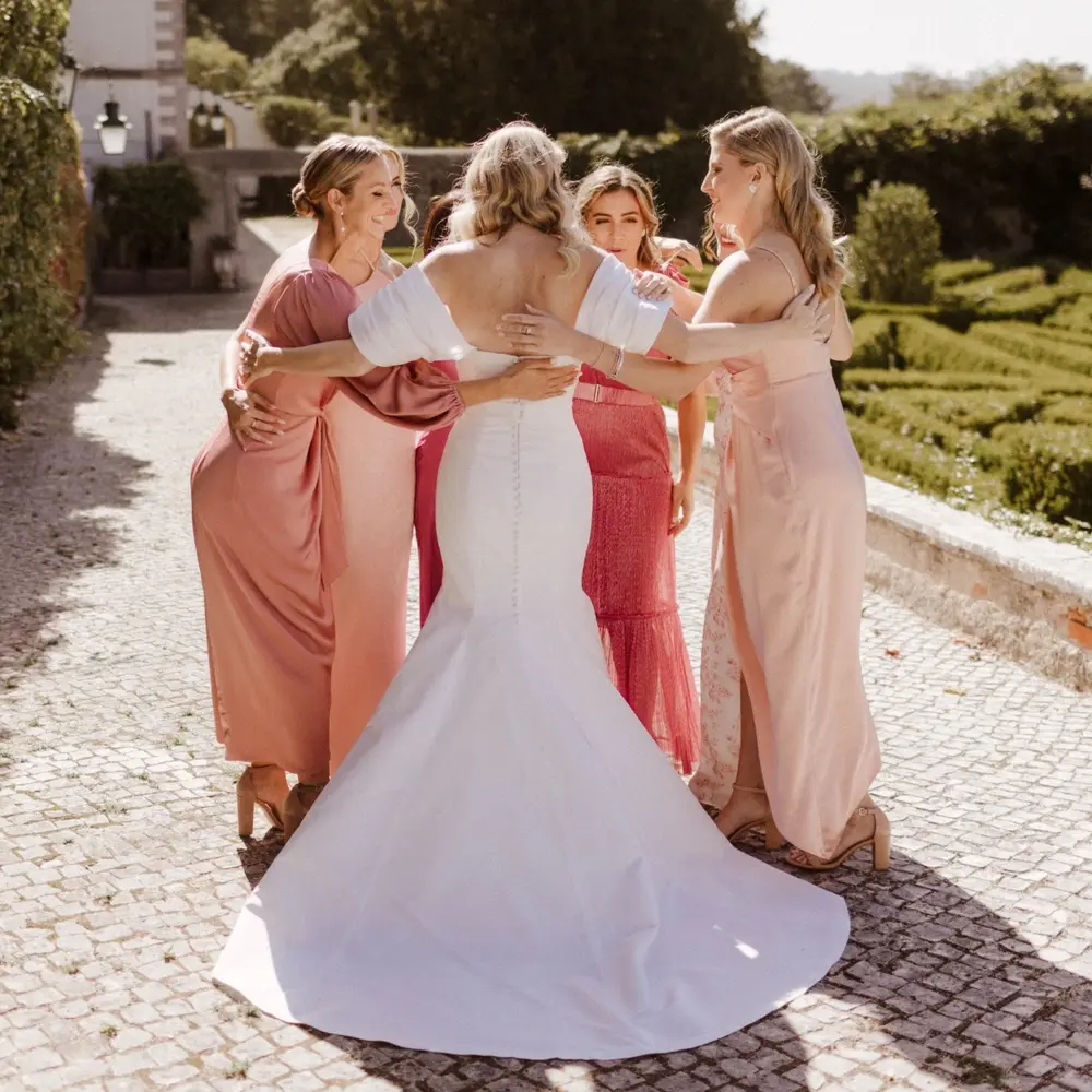 Bride in fishtail gown embraces three bridesmaids in pink dresses on a sunlit cobbled terrace