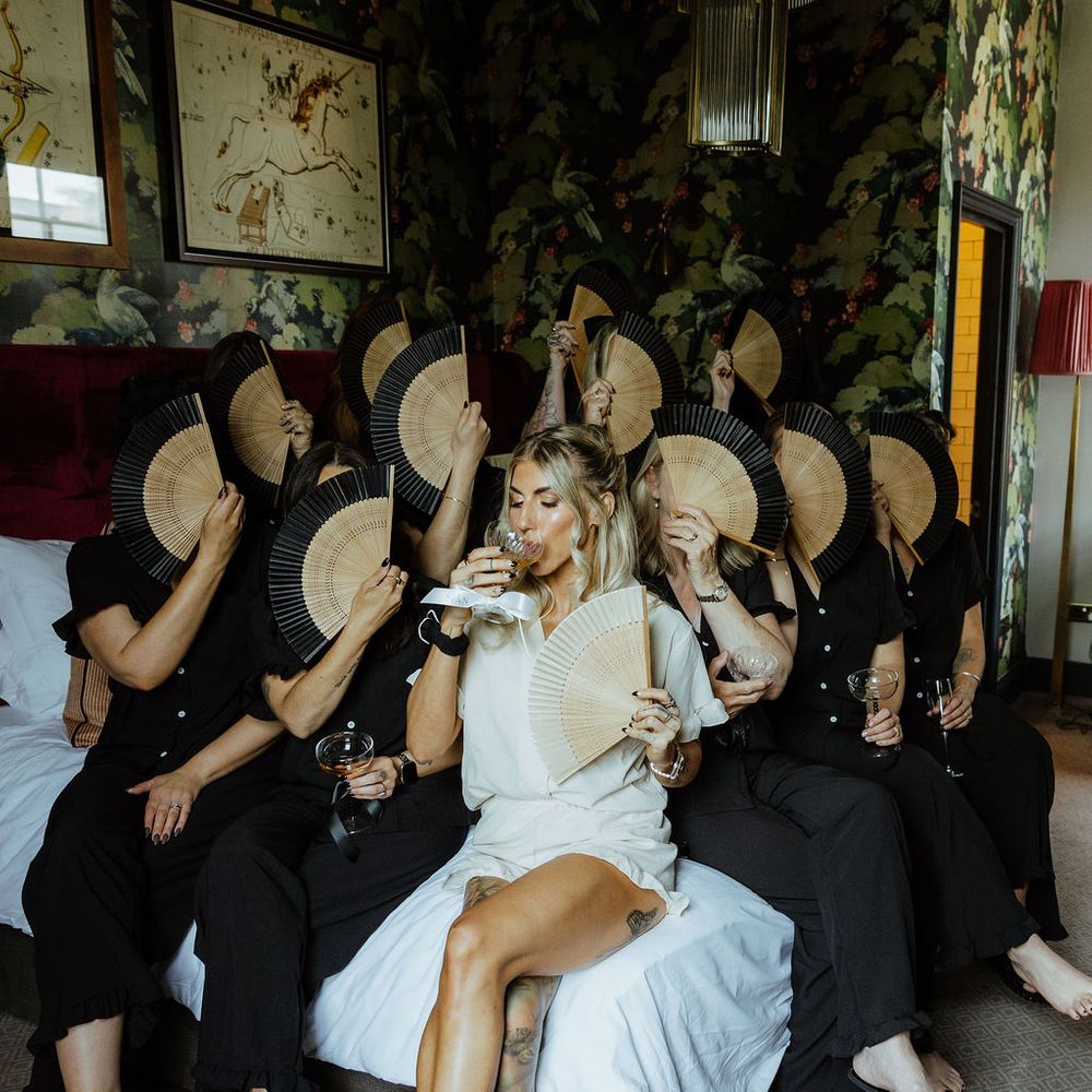 Bride sips a drink surrounded by bridesmaids in black holding wooden fans over their faces