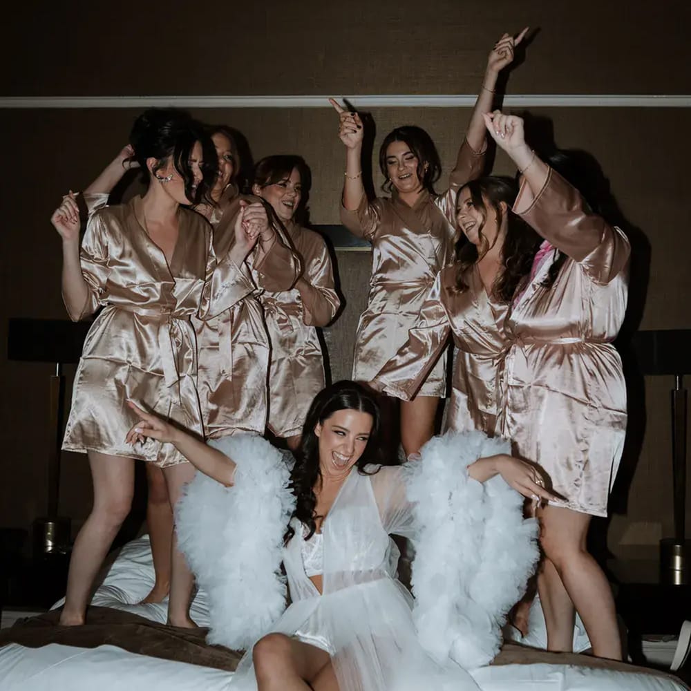 Bride in tulle robe laughing on a bed as bridesmaids in champagne satin robes dance behind her