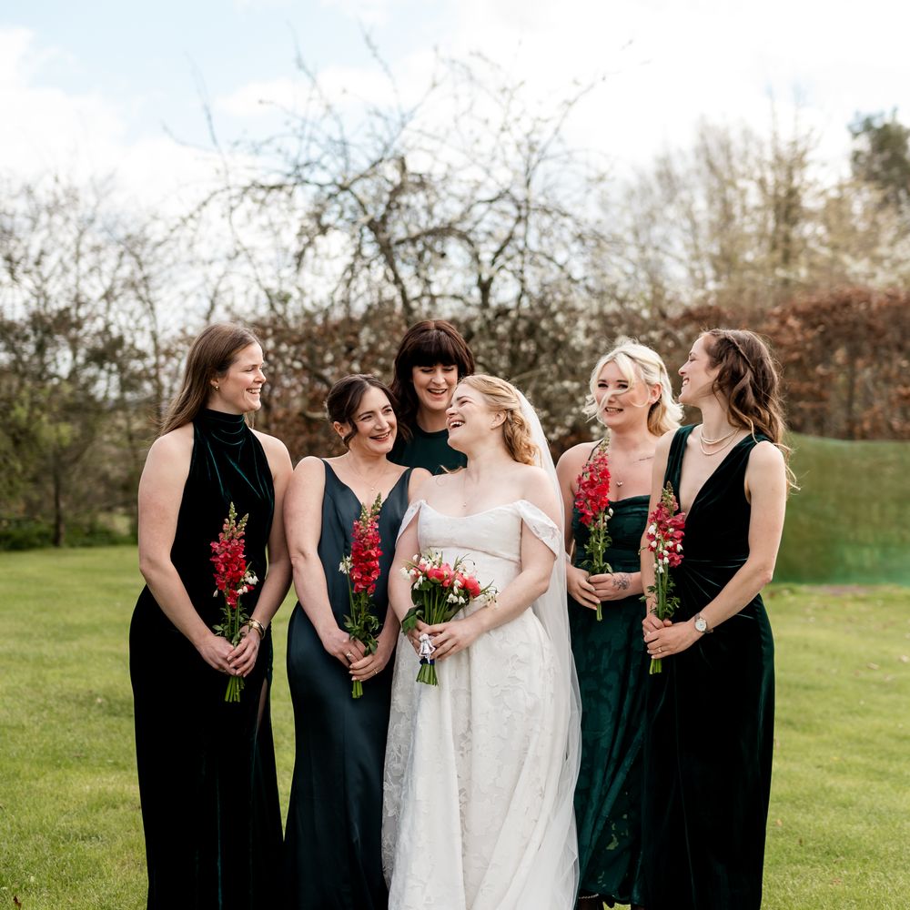 Bride in lace gown laughing with bridesmaids in green velvet dresses holding pink blooms