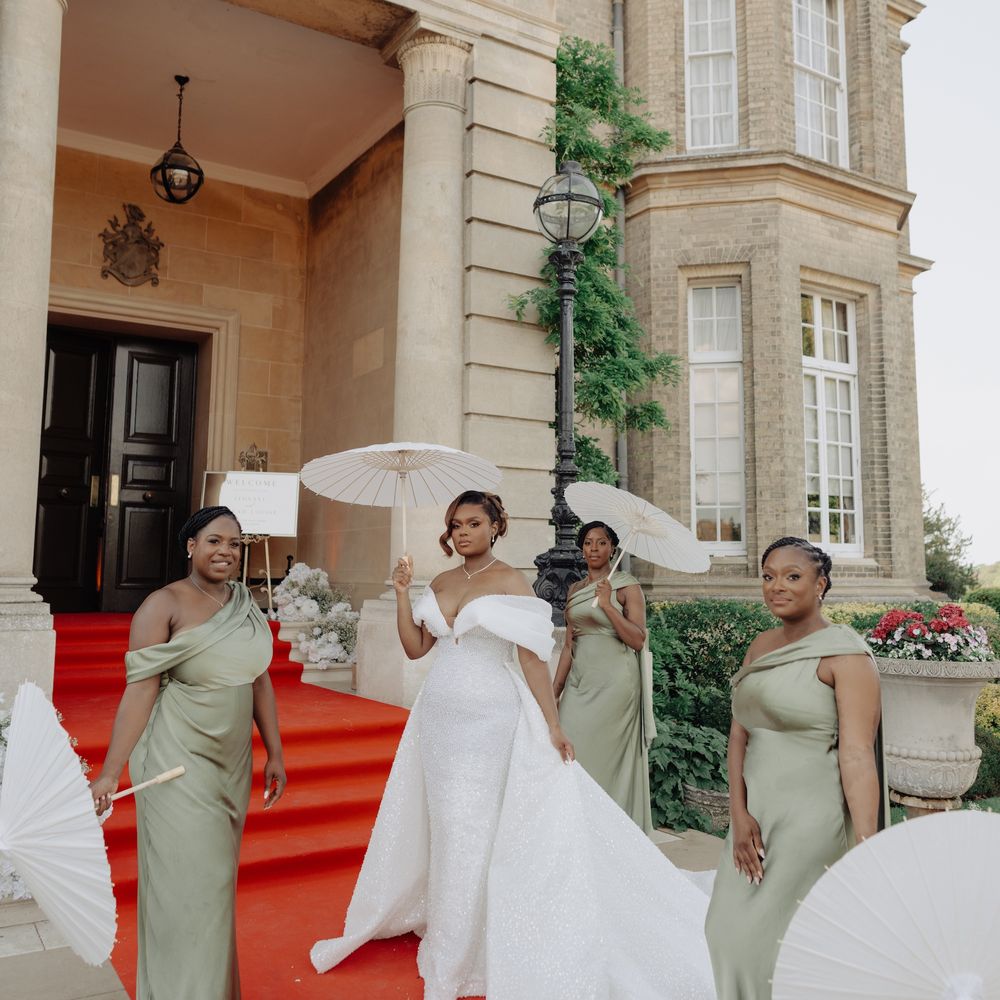 Bride and bridesmaids in sage green gowns holding white parasols outside a grand manor house