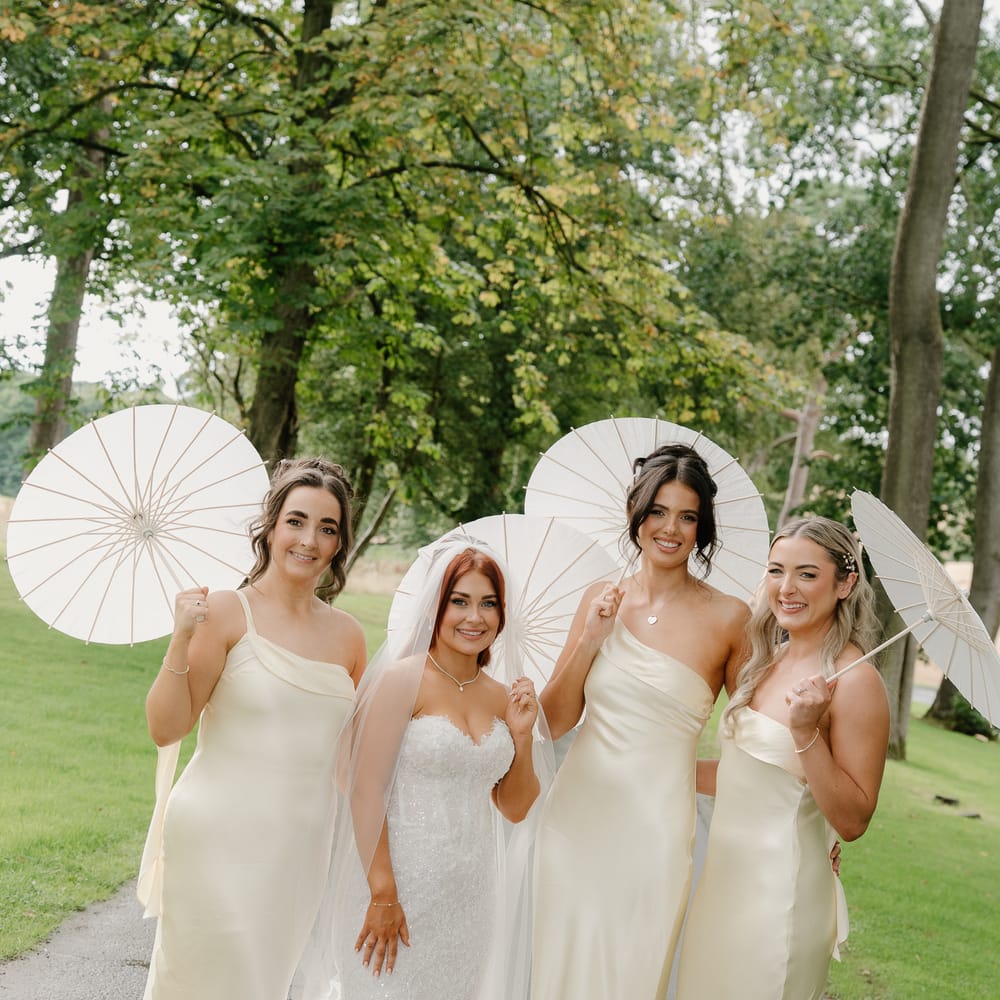 Bride with three bridesmaids in buttercup silk gowns holding white paper parasols on a tree-lined path
