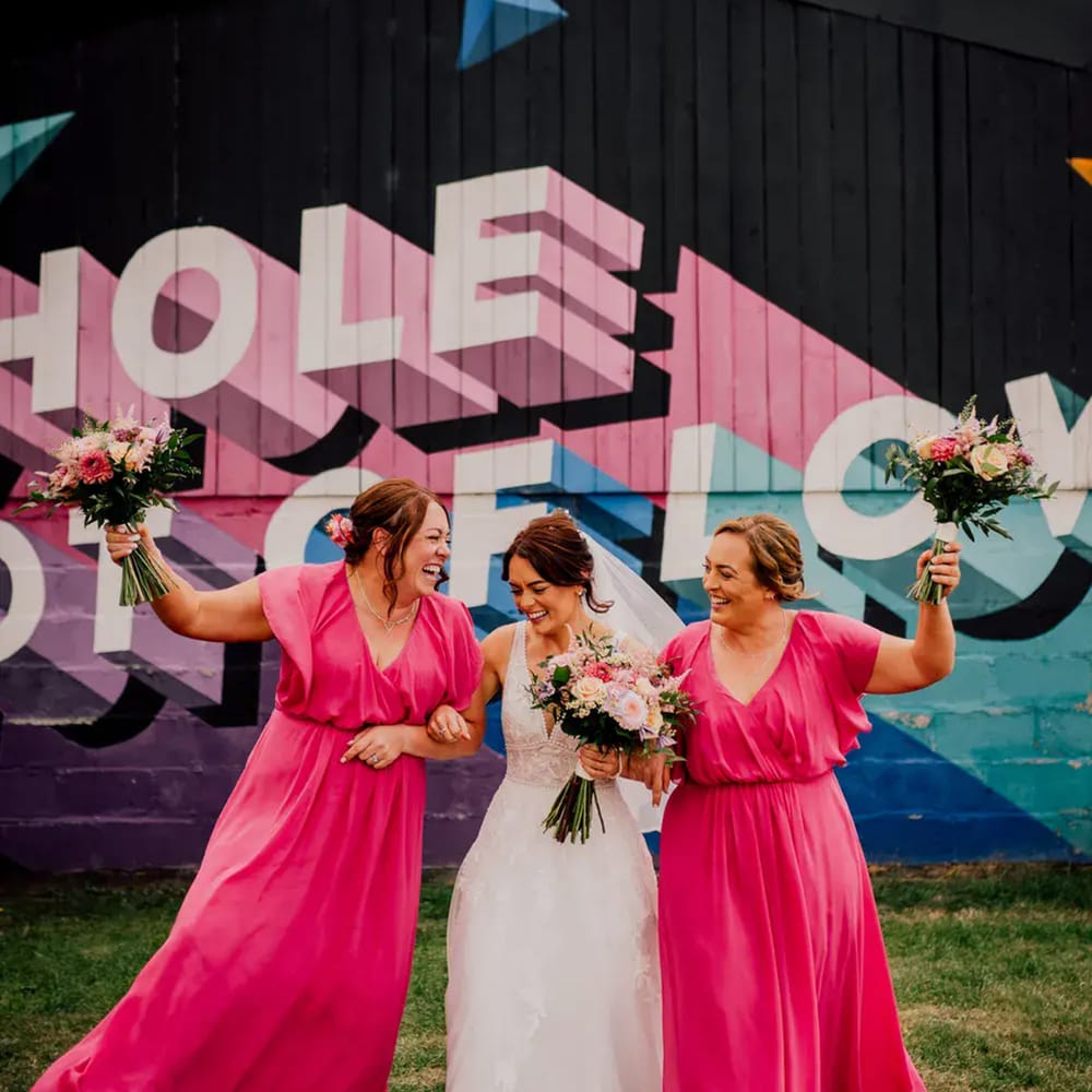 Bride walks arm in arm with two bridesmaids in hot pink gowns against a colourful mural