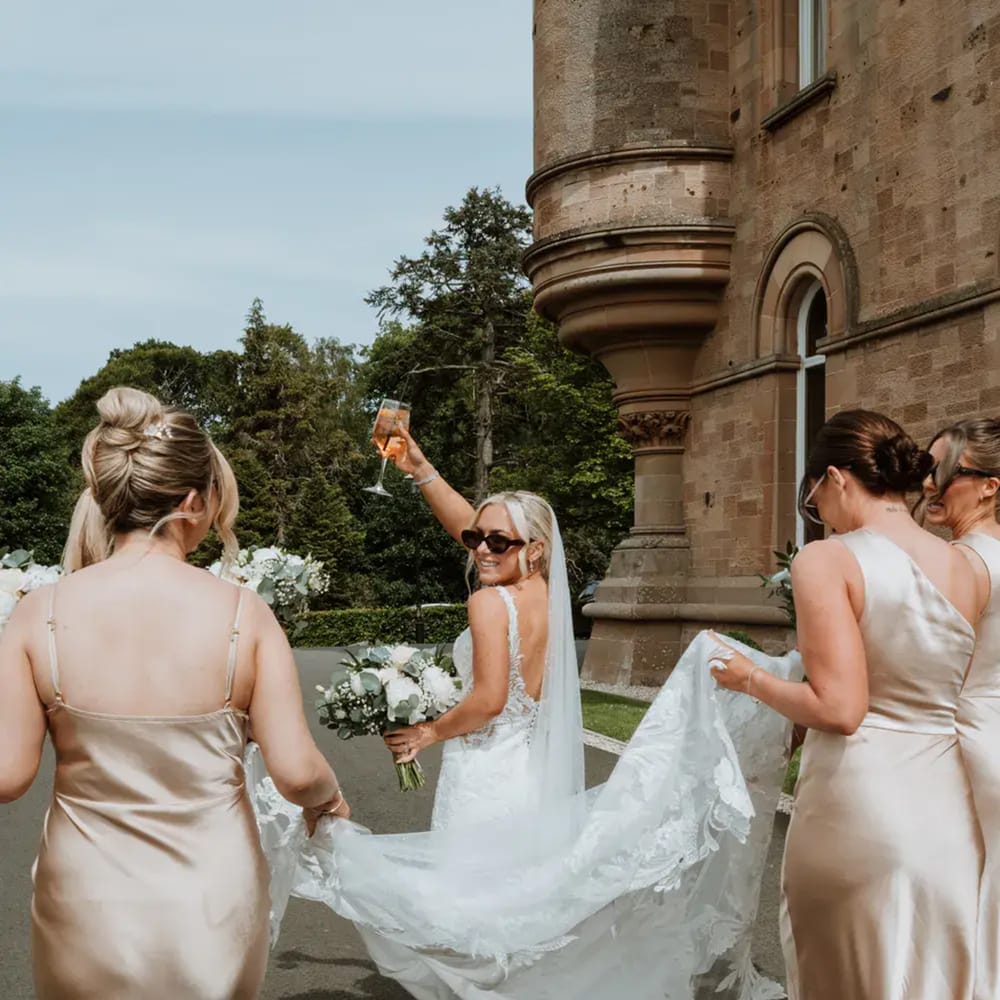 Bride raising a glass with sunglasses on while bridesmaids in champagne satin carry her veil
