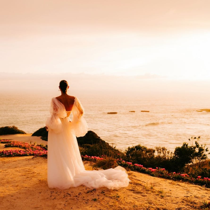 Bride in a flowing gown gazes out to sea from a clifftop at golden hour