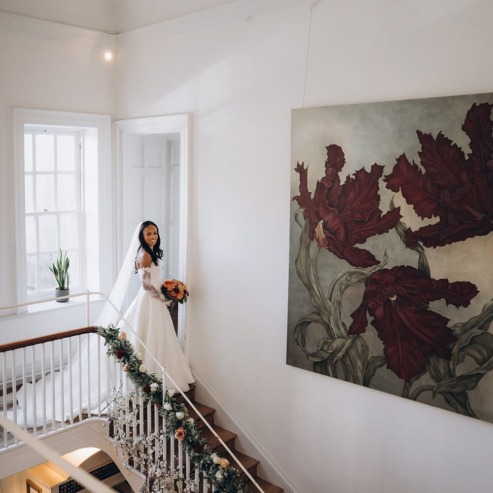 Bride in off-shoulder gown holding autumnal bouquet descends a floral-garlanded staircase