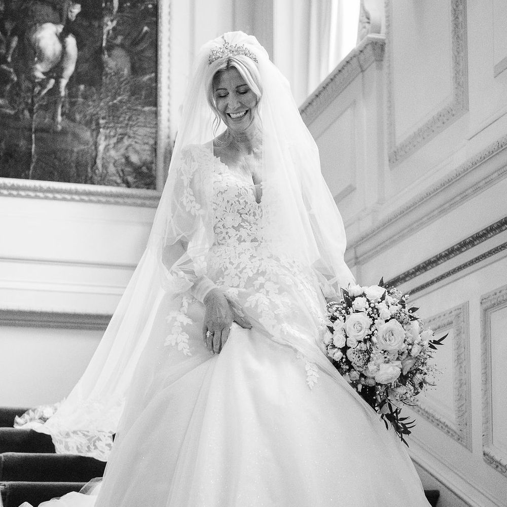 Smiling bride in lace ball gown and tiara descending a grand staircase with rose bouquet
