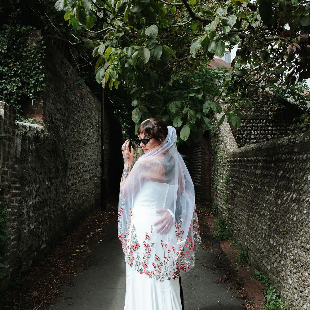Bride in sunglasses and a floral-embroidered veil glancing back along a walled garden path